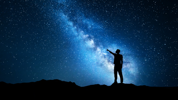Silhouette of a standing young man pointing finger in night starry sky on the background of blue Milky Way. Colorful night landscape. Beautiful Universe, travel background with sky full of stars