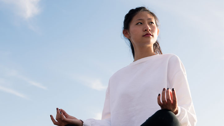 Asian girl in meditation pose looking to her left with blue sky with a few clouds in the background