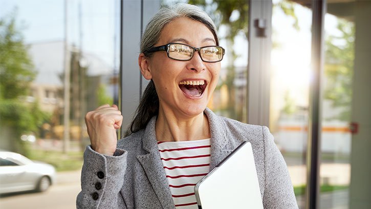 older woman holding laptop outside building fist pumping