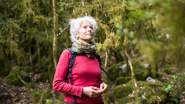older woman with a red shirt and a back pack looking up as she walks through the woods