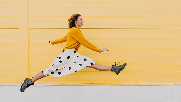 brown-haired woman in a polka dot skirt and golden sweater leaping in front of a yellow wall.