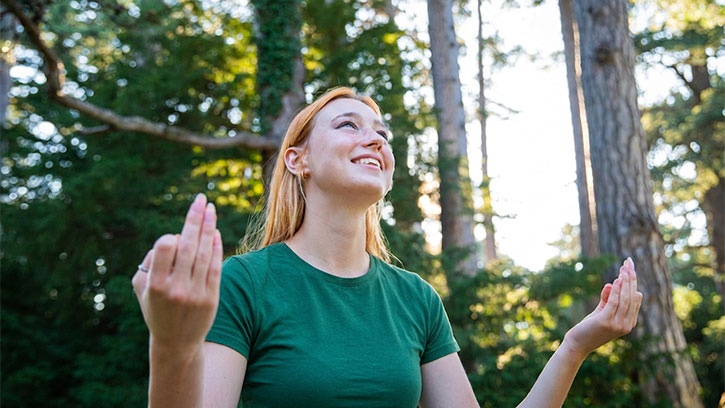 A smiling young woman with long red hair poses in a lush green forest. She has raised her hands. She is wearing a dark green top. The sun filters through the canopy of tall trees behind her.