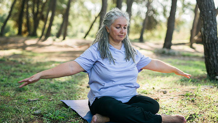 A senior woman with gray hair and a light purple shirt sits cross-legged on a blue yoga mat in a sunny wooded area, with arms stretched out to each side.
